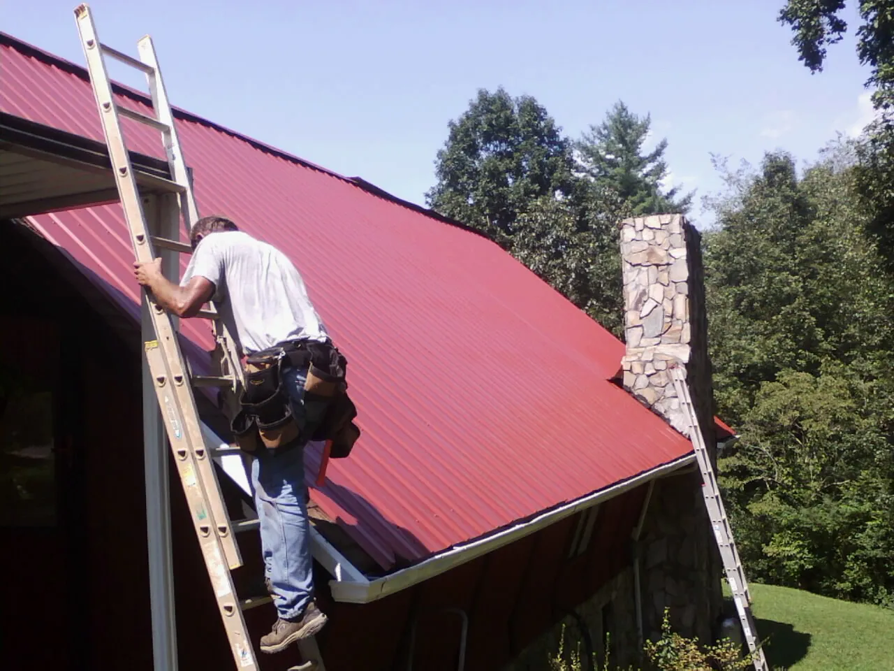 Roofer on a ladder beside a red metal roof and stone chimney — Soffit & Fascia Repair by Roofing Brothers in Colman