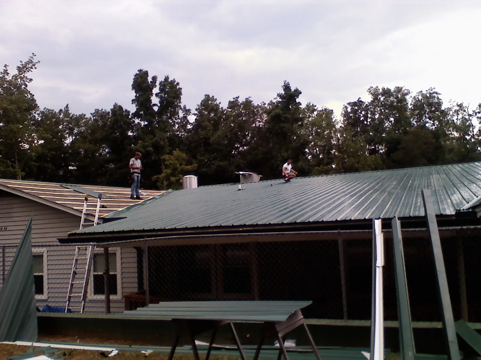 Two roofers installing green metal panels above a covered porch in Colman